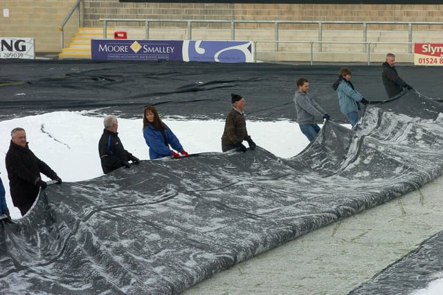 Morecambe fans, grounds staff and club officials help get snow of the covers protecting the pitch at the Globe Arena on Boxing Day in preparation for the fixtures against Aldershot and Accrington Stanley in 2003