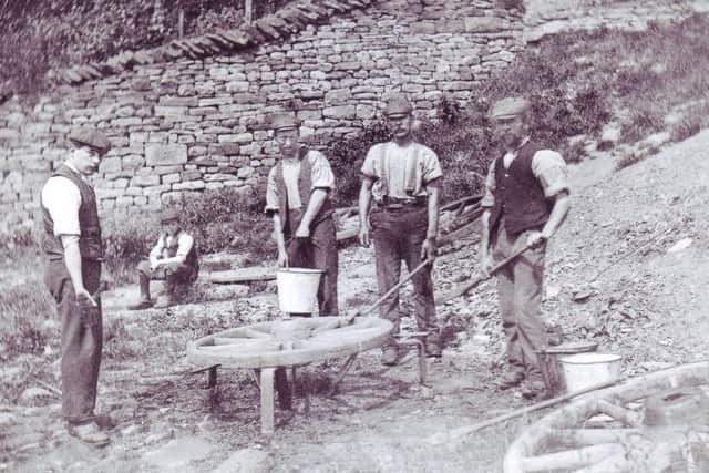 On the bank of the River Roeburn, just down from the smithy that was situated at the top of the bank above. David Travis (second from right) and his helper are holding iron dogs. These were used for carrying the red hot wheel rim from the fire, then placing it over the wheel. Water was then thrown over the iron rim to shrink it tight on to the wooden wheel.
After the wheels were hooped they would go back to the carpenters workshop to be painted with three coats of red lead paint. At this time there were steps down from the top of Smithy Brow to the rivers edge with stepping stones across the river. All this part of the river bank was destroyed by the Wray flood on August 8 1967.